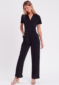 Combinaison noire à manches courtes, décolleté en V, taille cintrée et pantalon large. Tissu lisse et accent de bouton à la taille.