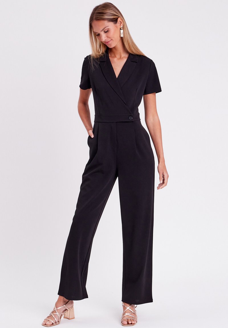 Combinaison noire à manches courtes, décolleté en V, taille cintrée et pantalon large. Tissu lisse et accent de bouton à la taille.