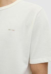 Tricou alb cu mânecă scurtă din material texturat, cu decolteu rotund și un mic detaliu metallic cu logo pe partea superioară stângă a pieptului.