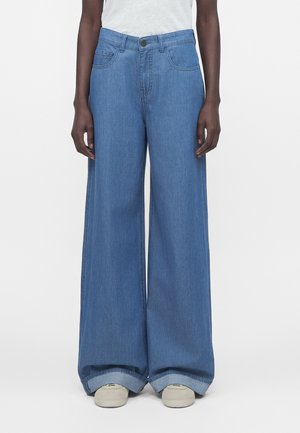 Person iført blå vidde-ben cowboybukser med store opslag og hvide sneakers, stående mod en ensfarvet hvid baggrund.