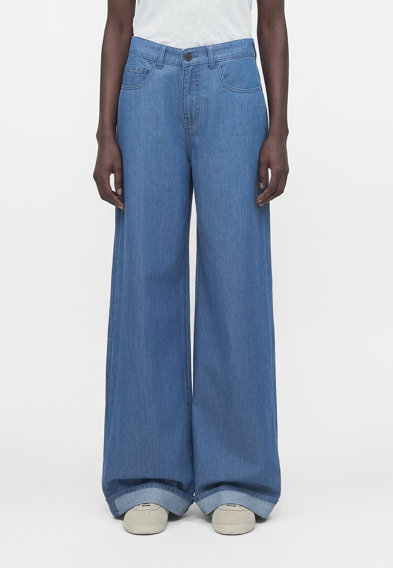 Άτομο που φοράει μπλε τζιν με φαρδιά μπατζάκια και μεγάλες γυρίσματα, με λευκά αθλητικά παπούτσια, στέκεται μπροστά σε απλό λευκό φόντο.