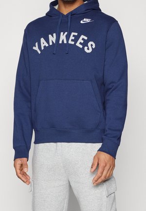 Homme portant un sweat à capuche Nike bleu marine avec le texte « YANKEES » et un pantalon cargo gris clair, debout devant un fond uni.
