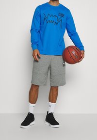 Henkilö, joka pitää oikeassa kädessään Nike-koripalloa ja on pukeutunut siniseen pitkähihaiseen paitaan, harmaisiin shortseihin, valkoisiin Puma-sukkiin ja mustiin lenkkareihin.