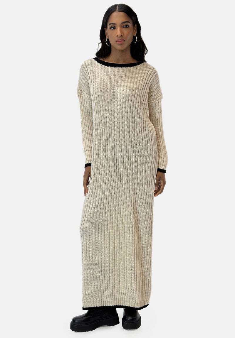 Strick-Maxikleid in Creme mit geripptem Textur und schwarzem Besatz am Ausschnitt und an den Ärmeln, getragen mit klobigen schwarzen Stiefeln.