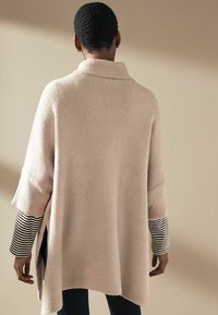 Pull beige surdimensionné avec un col montant, des poignets côtelés et des fentes sur les côtés, superposé à une chemise à manches longues rayée noir et blanc.