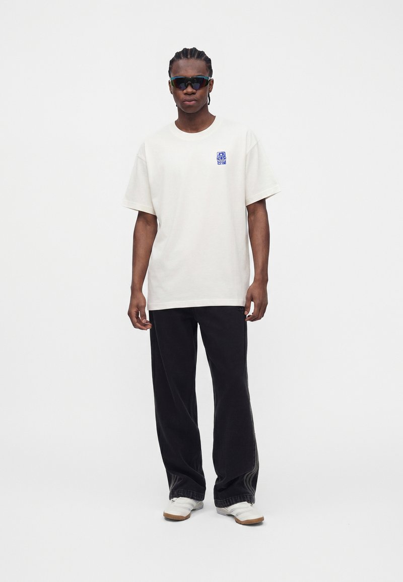 Personne portant un t-shirt blanc oversize avec un petit motif bleu, un pantalon large noir, des baskets blanches et des lunettes de soleil foncées, debout devant un fond uni.