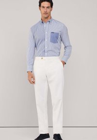 Camicia a righe blu e bianche con taschino sul petto, abbinata a pantaloni bianchi e scarpe scure. Tessuto in cotone morbido, vestibilità su misura.