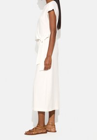 Biała sukienka do kostek z krótkimi rękawami, z paskiem wiązanym w talii, noszona z brązowymi sandałami na paski. Gładka tekstura materiału, minimalistyczny design.