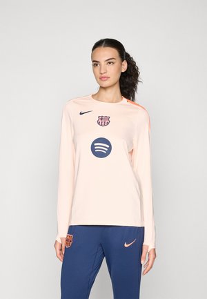 Chemise de sport à manches longues, de couleur pêche clair, fabriquée en tissu lisse ; présente un logo marine et un graphisme Spotify à l'avant, avec des accents orange.
