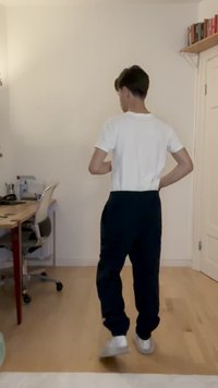Hvit kortermet skjorte og marineblå joggebukser, med en avslappet passform, iført hvite joggesko. Trelastgulv, nøytral vegg i bakgrunnen.