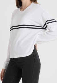 Vit långärmad topp med ribbad struktur, som har ett horisontellt svart rand över bröstet och en rundad nederkant, kombinerad med svarta jeans.