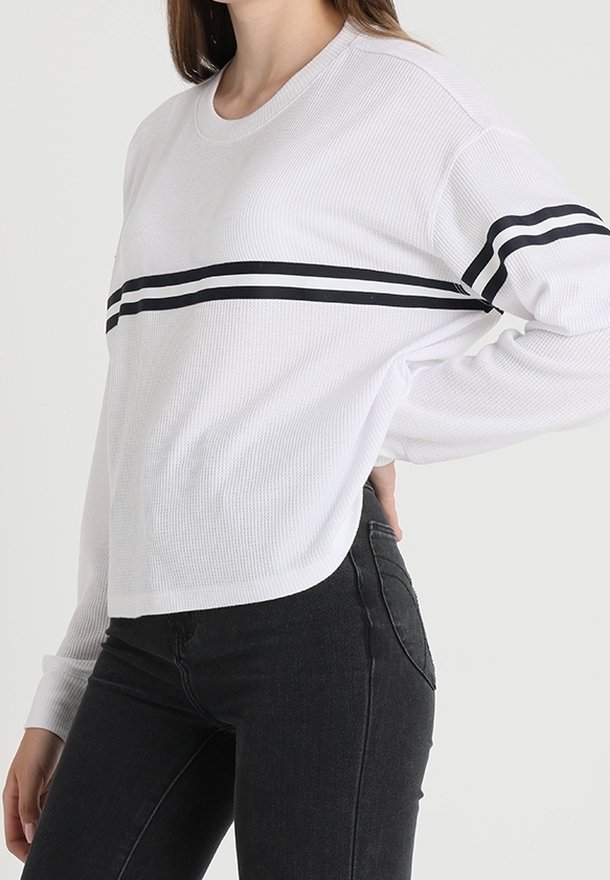 Vit långärmad topp med ribbad struktur, som har ett horisontellt svart rand över bröstet och en rundad nederkant, kombinerad med svarta jeans.
