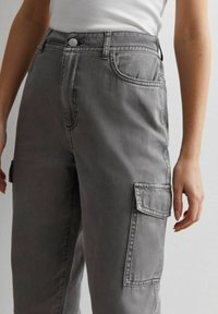 Person, der hoch taillierte graue Cargo-Hosen mit Front- und Seitentaschen trägt, kombiniert mit einem weißen, gerippten, ärmellosen Oberteil.