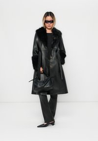 Sort læderjakke med faux fur krave og manchetter, parret med tætsiddende sorte bukser og en struktureret sort håndtaske. Model iført mørke solbriller.