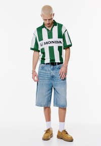 Groen en wit gestreepte polo met "HONDA" logo, gecombineerd met lichtblauwe denim shorts. Bruine schoenen passen bij witte sokken.