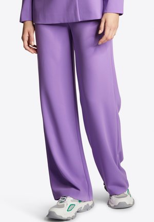 Femme portant un pantalon ample lavande et des baskets blanches avec des détails verts, debout devant un fond blanc uni.