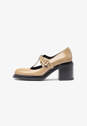 Zapato Mary Jane de charol beige con tira en T, detalle de hebilla dorada y un tacón grueso negro. Superficie lisa, punta redondeada.