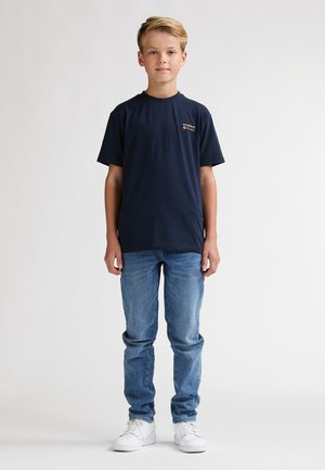 Camiseta azul marino con cuello redondo, mangas cortas y un pequeño logo en el pecho, combinada con vaqueros azul claro y zapatillas blancas.