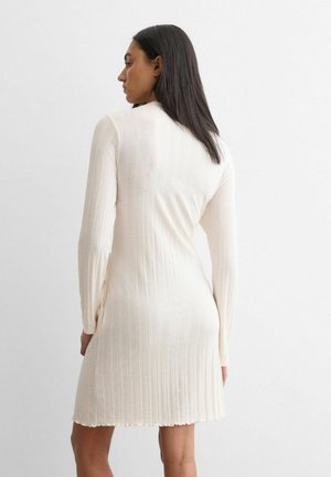 Kvinde med lige mørkt hår iført en langærmet, cremefarvet ribstrikket kjole, stående mod en ensfarvet hvid baggrund, vendt væk.