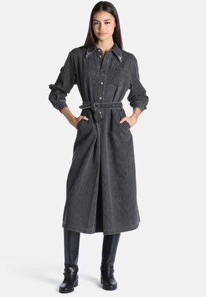 Femme portant une longue robe en jean noire avec une ceinture, un col orné, et des bottes noires montantes, debout les mains dans les poches.