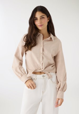 Giovane donna con lunghi capelli scuri, che indossa una camicia beige con bottoni annodata in vita e pantaloni bianchi a vita alta, su uno sfondo chiaro.