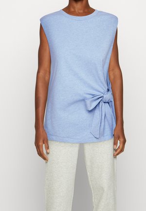 Sleevetøs lyseblå top med sidesløjfedetalje, iført med lysegrå bukser på en person, der står foran en ensfarvet baggrund.