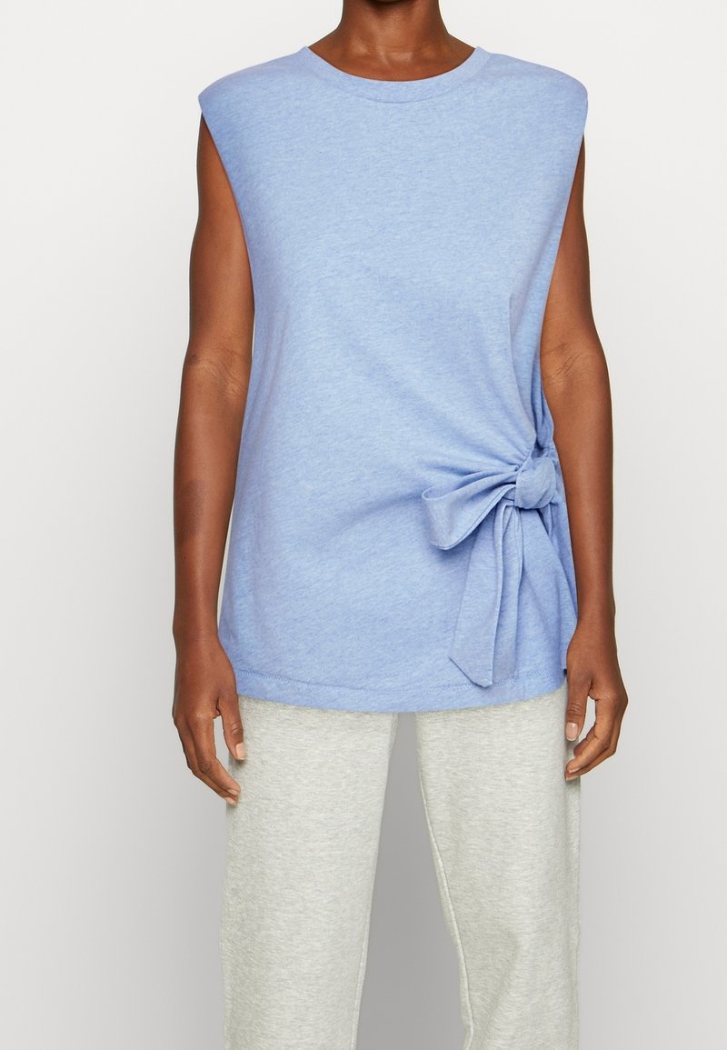 Top sans manches de couleur bleu clair avec un détail de nœud sur le côté, porté avec un pantalon gris clair par une personne se tenant devant un fond uni.