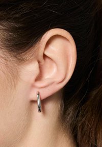 Srebrna kolczyk w kształcie obręczy umieszczona na małżowinie usznej, z wypolerowaną, gładką powierzchnią i okrągłym, wydłużonym kształtem. Zbliżenie.