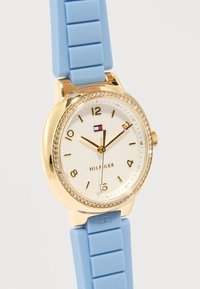 Ρολόι με στρογγυλό καντράν σε χρυσό τόνο, στεφάνη με κρύσταλλα, λευκό υφασμένο καντράν, χρυσούς αριθμούς και δείκτες, μπλε λουράκι σιλικόνης και λογότυπο Hilfiger στο καντράν.