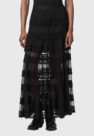 Jupe maxi en dentelle noire à étages avec des panneaux transparents portée avec des bottes noires, montrant le bas du corps d'une personne sur fond gris.