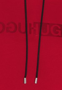 Sweat à capuche rouge avec cordons noirs, présentant un grand logo embossé sur le devant et des embouts métalliques argentés sur les cordons. Texture douce.