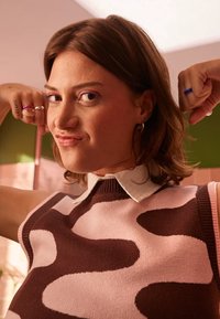 Gebreide mouwloze top met een bruin en roze abstract patroon, geribbelde halslijn en een witte met kraag shirt eronder, met minimale accessoires.