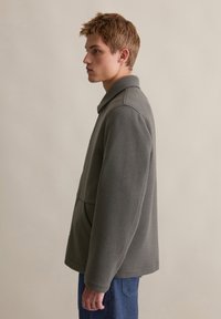 Grijze fleecejack met een knoopsluiting aan de voorkant, een opstaande kraag en zijzakken, weergegeven op een model in profiel tegen een neutrale achtergrond.