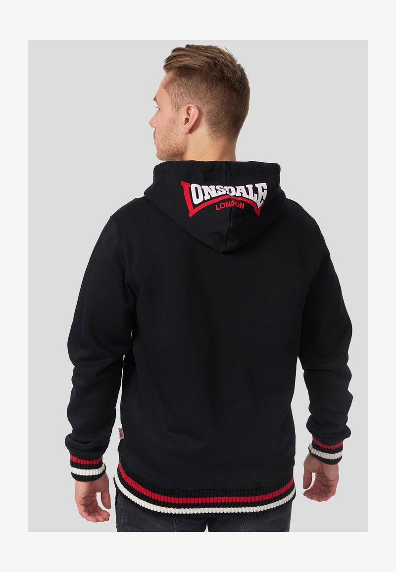 Mees, kes seisab seljaga, kannab musta kapuutsiga pusa, millel on kapuutsil valge ja punane "Lonsdale London" logo, punase ja valge triibuliste varruka- ja alumiste äärega.