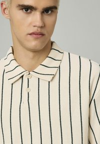 Polo shirt beige rayé avec une texture douce, présentant des rayures verticales vert foncé, un col classique et deux boutons sur le devant.