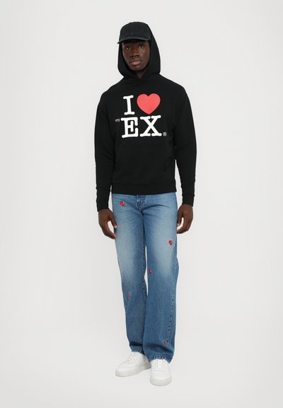 Sort hættetrøje med hvidt og rødt tekst; blå jeans med røde hjerteaccenter; hvide sneakers; model stående mod en ensfarvet baggrund.