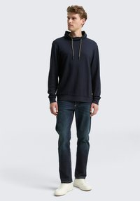 Marineblauwe hoodie met een funnel neck en gouden trekkoordjes, gecombineerd met donkere jeans en witte sneakers. Glad textuur met een slim fit ontwerp.