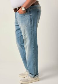 Jeans de mezclilla decolorados y desgastados en azul claro, con un ajuste relajado, que presentan desgarros y un aspecto desvanecido. Llevados con un cinturón marrón y zapatillas blancas.