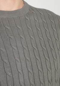 Grå stickad tröja med en trefärgad kabelmönster, rund halsringning och ribbade ärmslut. Materialet verkar mjukt och bekvämt.