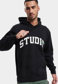 Svart hettegenser med hvit "STUDIO"-skrift, kengurulomme, snorhette og ribbestrikkede cuffs. Materialet ser mykt og teksturert ut.