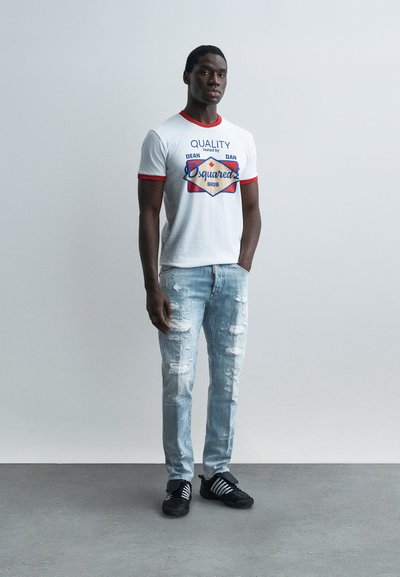 Homme debout les mains dans les poches, portant un t-shirt blanc à motif avec une bordure rouge, un jean clair légèrement usé, et des baskets noires à rayures blanches.