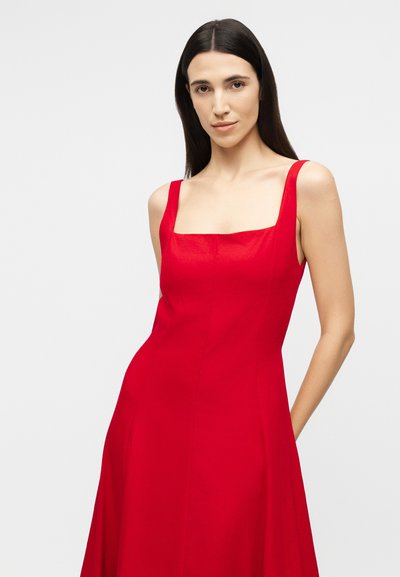 Vrouw met lang donker haar, een mouwloze rode jurk met een vierkante halslijn dragend, staand tegen een effen lichtgrijze achtergrond.