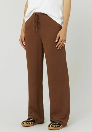 Pantalons légers, larges et marron avec une taille à cordon ; associés à des ballerines imprimées léopard noires et beiges. Texture lisse.