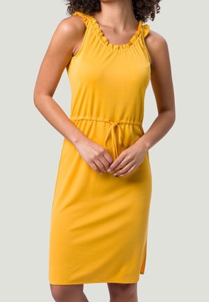 Femme portant une robe jaune sans manches avec un col à volants et une taille à cordon, debout, les mains jointes devant elle.