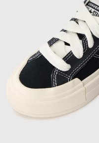 Black canvas sneaker with cream rubber sole, featuring white laces and contrasting stitching details. Rounded toe and padded collar design.