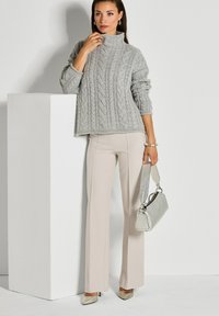 Vrouw in een grijze kabelgebreide trui en beige broek, met een zilveren handtas, staande naast een witte rechthoekige sokkel.