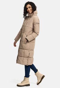 Beige puffer coat with a high collar, featuring a belted waist, stitched design, and snap buttons, paired with beige lace-up boots and denim.
