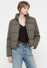 Ung person med kort hår iført en olivengrøn pufferjakke, sort V-hals top, lyseblå jeans og sort bælte med guldbuckle.