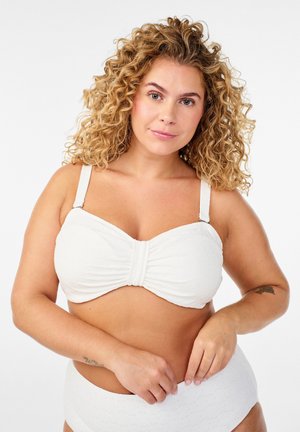 Krøllet kvinde med lys hud iført en hvid tekstureret bikini, der justerer bikinitrusserne mod en ensartet hvid baggrund.