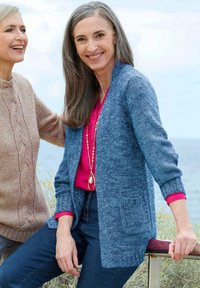 Zwei lächelnde ältere Frauen im Freien am Wasser, eine in einem blauen Cardigan und pinkem Shirt, die sich an einem Geländer lehnt, die andere in einem beigen Pullover neben ihr.